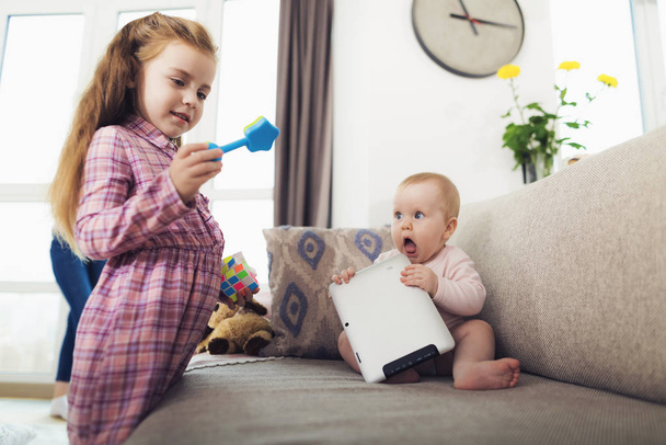 Krásná dívka si hraje s malou sestřičku uvnitř. Roztomilý dlouhosrstý dítě drží modré hračky a visí ven s mladší sourozenec udržet Tablet v rukou sedí na gauči v obýváku moderní. - Fotografie, Obrázek