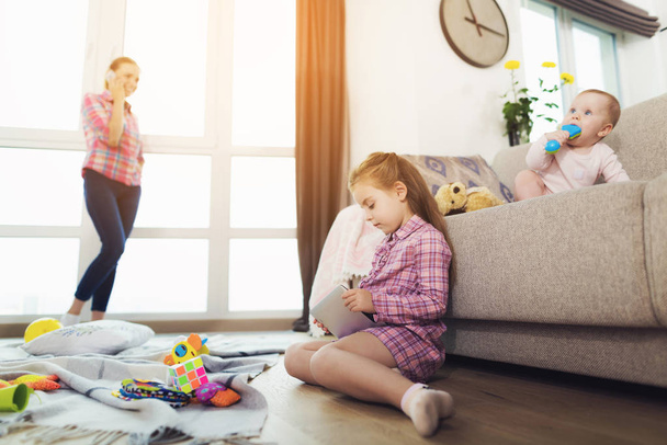 Children Playing in Room while Mom Talking Phone. Mother Standing near Large Panoramic Window in Living Room. Baby Sitting on Couch Keeping Toy in Mouth and Older Child Using Tablet on Floor - Photo, image