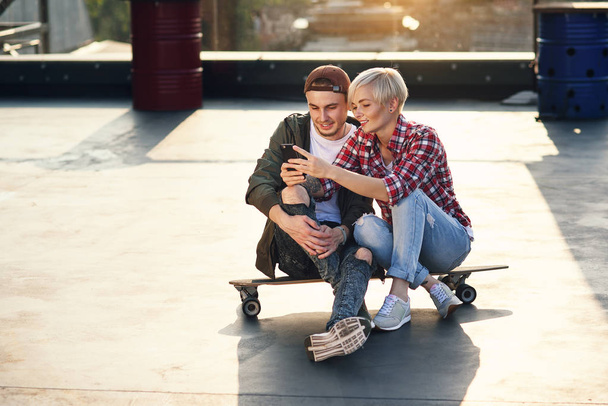 Handsome stylish couple sitting on longboard skateboard on the rooftop playground of industrial building and using smartphone. Modern urban active lifestyle concept. - Фото, изображение