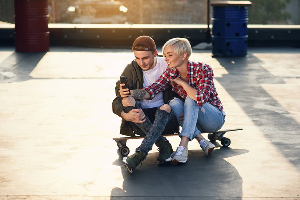 Handsome stylish couple sitting on longboard skateboard on the rooftop playground of industrial building and using smartphone. Modern urban active lifestyle concept. - Foto, immagini