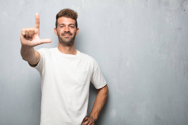 young handsome man gesturing "loser" with fingers mockingly, with a happy, proud, and self-satisfied look. - Fotoğraf, Görsel