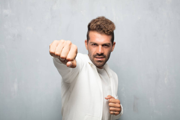young handsome man with an angry, aggressive and menacing pose, ready for the fight, showing fists furiously and belligerently. - Foto, afbeelding