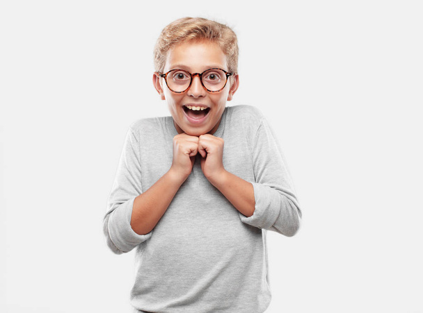 young blonde handsome boy with a happy surprised expression, eyes wide open and broad smile, with both hands clenched under chin showing shock. - Photo, Image