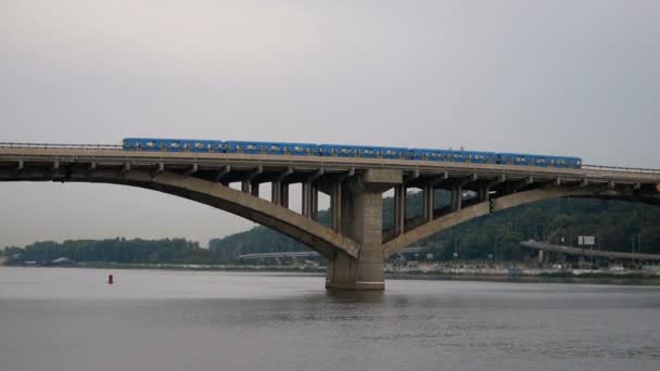 Zug fährt über die Brücke gegen Himmel und Wald. City-U-Bahn links außen. Öffentliche Verkehrsmittel in Bewegung. Autos fahren über die Brücke über den Fluss. - Filmmaterial, Video