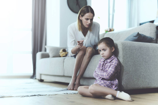 Upset Girl Sitting on Floor while Mother Shouting. Little Pretty Child near Light Couch Taking Displeasure when Woman Yelling with Spreaded Hands. Family Relationship Concept - 写真・画像