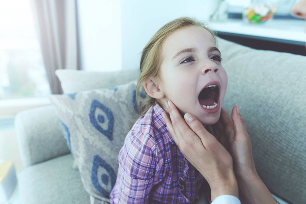 Sick Child Opens Mouth Widely and Showing Throat. Closeup of Female Doctor Hands Holding Gently Girl Neck. Caucasian Kid Sitting on Couch in Living Room. Child Healthcare Concept - Foto, afbeelding