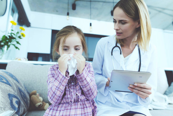Sick Kid Blowing Nose and Sits Near Female Doctor. Child Has Cold Coughs in Handkerchief Sitting on Couch at Home. Young Nurse in White Medical Gown Making Diagnosis. Child Healthcare Concept - Photo, image