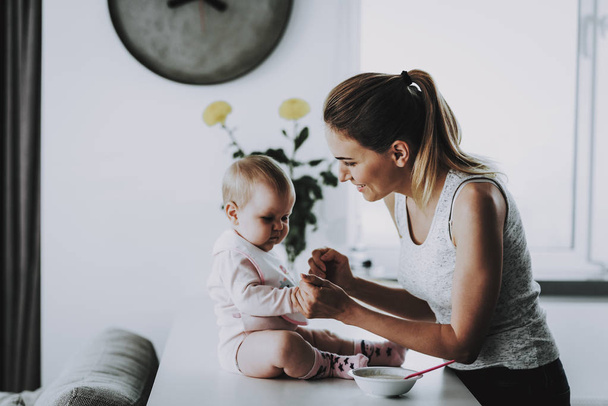 Happy Smiling Mother Feeds Adorable Kid at Home. In Bright Living Room Cute Hungry Toddler Having Breakfast with Mom while Sitting on Table near Bowl Motherhood and Childhood Concept - Photo, Image