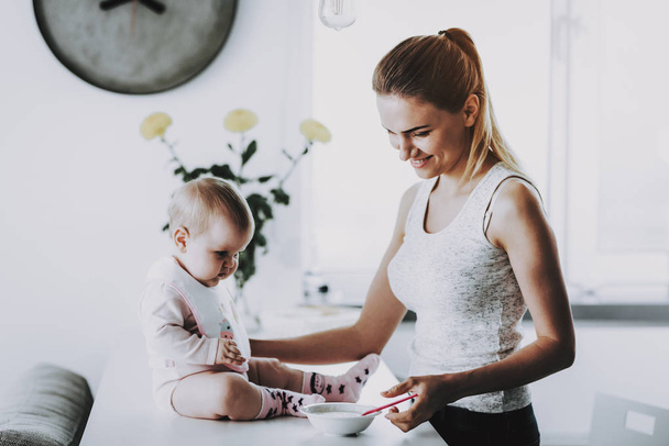 Happy Smiling Mother Feeds Adorable Kid at Home. In Bright Living Room Cute Hungry Toddler Having Breakfast with Mom while Sitting on Table near Bowl Motherhood and Childhood Concept - Foto, Imagen