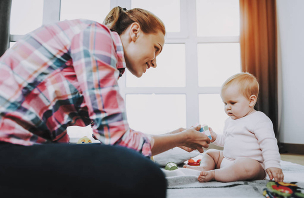 Young Mother Playing Toys with Cute Baby on Carpet. Smiling Mom Wears Casual Clothes Resting with Sweet Beautiful Caucasian Toddler near Large Panoramic Window in Living Room. Childhood Concept - Фото, изображение