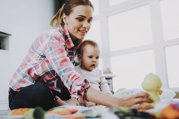Anya ül a gyermek a padló és a gazdaság baba. Fiatal mosolyogva anya visel alkalmi ruha játék játékok aranyos baba közelében nagy panoráma ablak a nappali padlón. - Fotó, kép