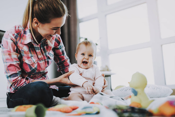 Mère est assise avec un enfant sur le sol et tenant la poupée. Jeune mère souriante porte des vêtements décontractés jouant des jouets avec bébé caucasien mignon sur le sol près de la grande fenêtre panoramique dans le salon
. - Photo, image