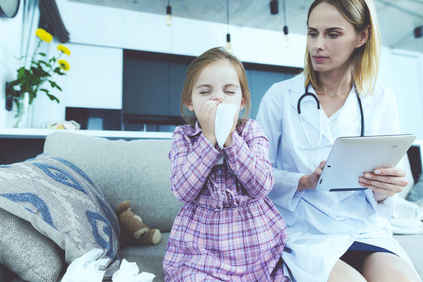 Kid Coughs in Handkerchief and Sits Near Doctor. Sick Child Has Cold Blowing Nose Sitting on Couch in Living Room at Home. Young Nurse in White Medical Gown Making Diagnosis. Child Healthcare Concept - 写真・画像