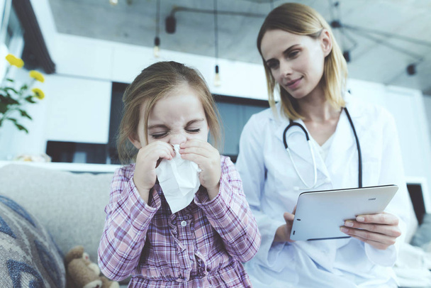 Sick Kid Blowing Nose and Sits Near Female Doctor. Child Has Cold Coughs in Handkerchief Sitting on Couch at Home. Young Nurse in White Medical Gown Making Diagnosis. Child Healthcare Concept - Foto, Imagen