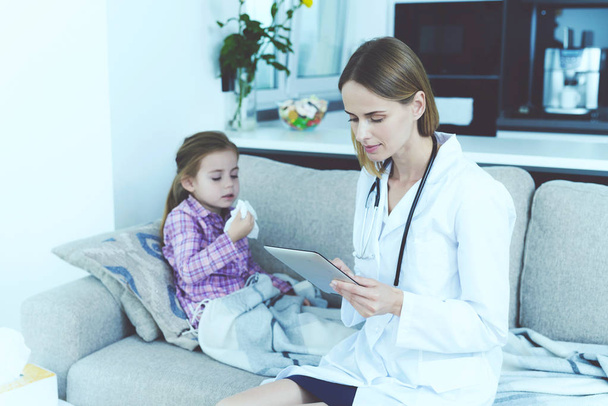 Young Female Doctor Hold Pad and Make Diagnosis. Pretty Little Ill Girl Having Cold Sitting on Couch with Nurse Wearing Stethoscope and White Medical Gown Looking at Tablet Screen. - Foto, imagen