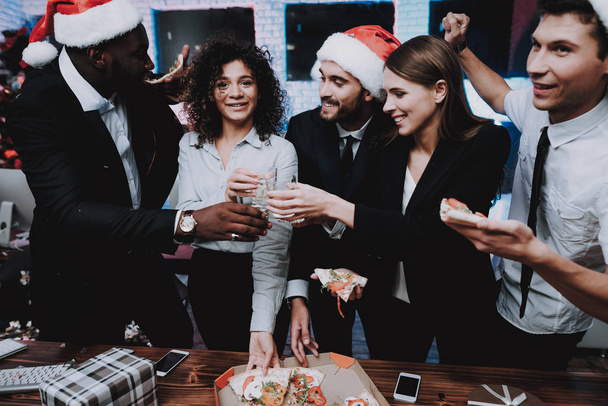 Young People Celebrating New Year in Modern Office. Christmas Tree in Office. Computer on Desk. People Have Fun. Indoor Party with Pizza. Celebrating of New Year. Man in Red Cap. Drinking Alkohol. - 写真・画像