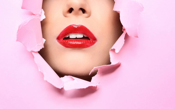 View of beautiful young woman with red lips through hole in color paper, closeup - Фото, изображение