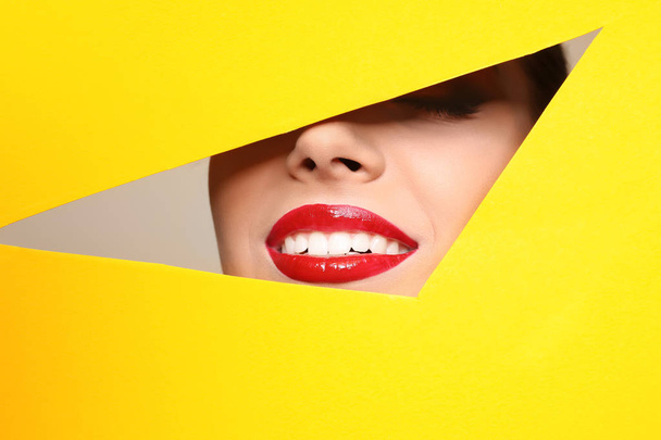 View of beautiful young woman with red lips through cutout in color paper, closeup - Foto, Imagem