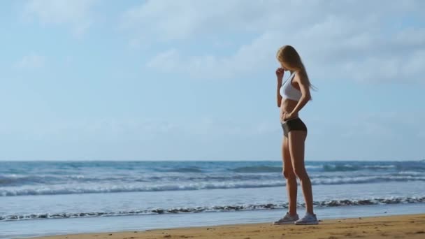 Κορίτσια τα πόδια σε αθλητικά ρούχα, στέκεται κοντά στον ωκεανό και να αγγίζει τα μαλλιά της σε αργή κίνηση. - Πλάνα, βίντεο