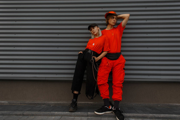 Young beautiful stylish couple models in fashion caps and trendy orange clothes are standing near the gray wall - Foto, Imagem
