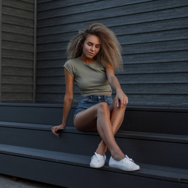 Sexy bella giovane donna modello con i capelli ricci in pantaloncini jeans casual e una t-shirt elegante con scarpe da ginnastica bianche alla moda seduto sui gradini della strada
 - Foto, immagini