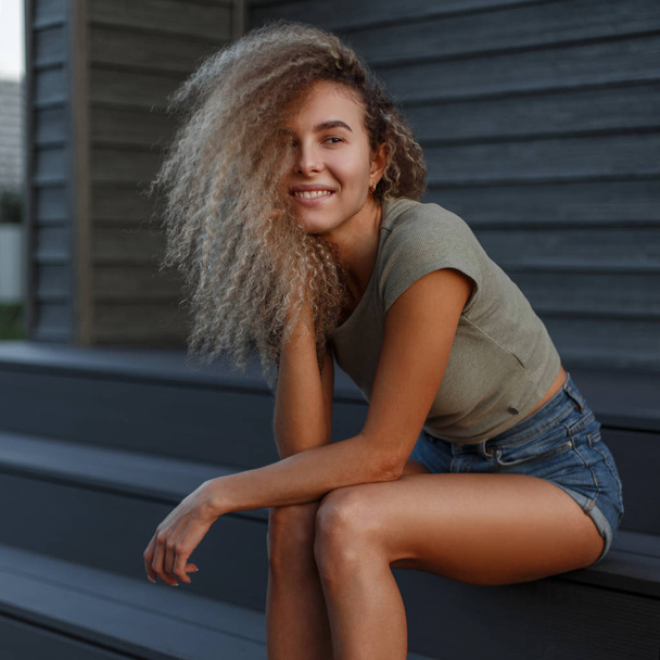 Happy krásná mladá roztomilá žena s úsměvem s kudrnatými vlasy v módní Jeans oblečení sedí na schodech - Fotografie, Obrázek