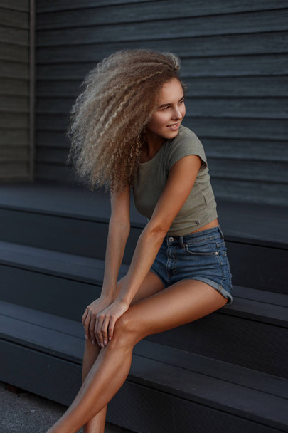 Happy beautiful funny curly woman with a smile in sexy jeans denim shorts sitting on the stairs - Фото, зображення