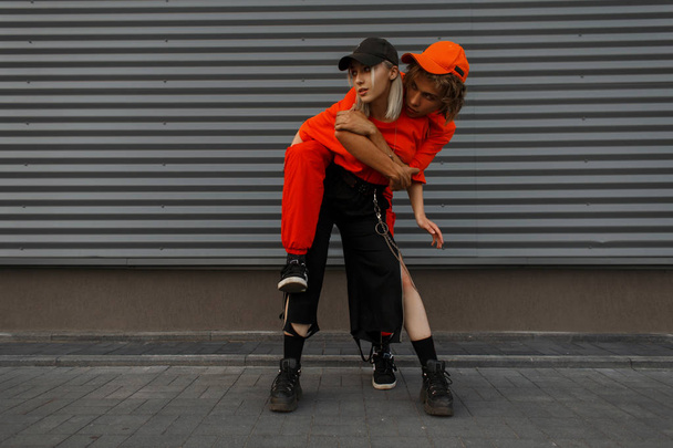 Красивая молодая смешная модная пара в стильной оранжевой одежде с модной кепкой рядом с металлической серой стеной на улице
 - Фото, изображение