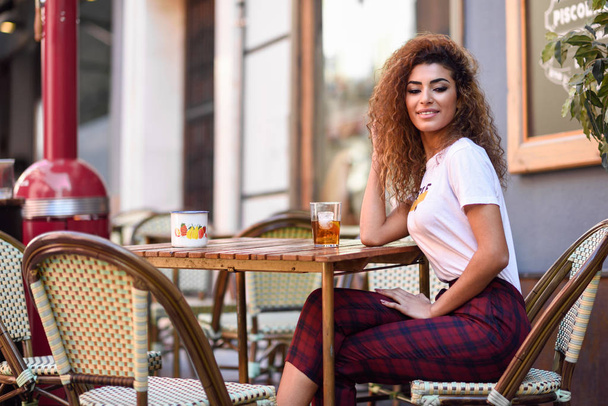 Young arabic woman smiling and sitting in an urban bar in the street. Arab girl in casual clothes drinking a soda outdoors. - Valokuva, kuva