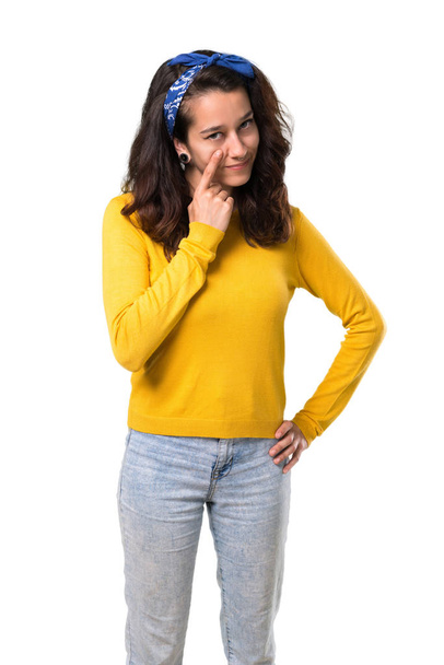 Young girl with yellow sweater and blue bandana on her head standing and looking to the front opening the eye with finger on isolated white background - Foto, Bild