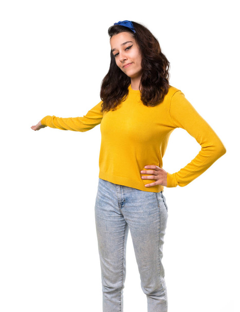 Young girl with yellow sweater and blue bandana on her head pointing back with the index finger presenting a product on isolated white background - Photo, image