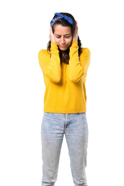 Young girl with yellow sweater and blue bandana on her head covering both ears with hands. Frustrated expression on isolated white background - Φωτογραφία, εικόνα