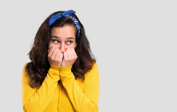 Young girl with yellow sweater and blue bandana on her head is a little bit nervous and scared putting hands to mouth on isolated grey background - Photo, Image