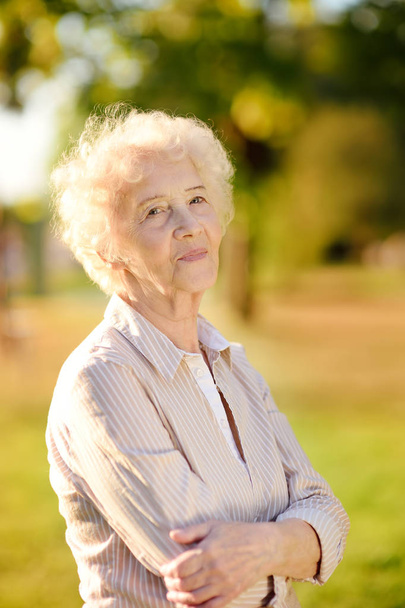 Outdoors portrait of beautiful smiling senior woman with curly white hair. Elderly lady walking in summer park. Active longevity concepts - Photo, Image