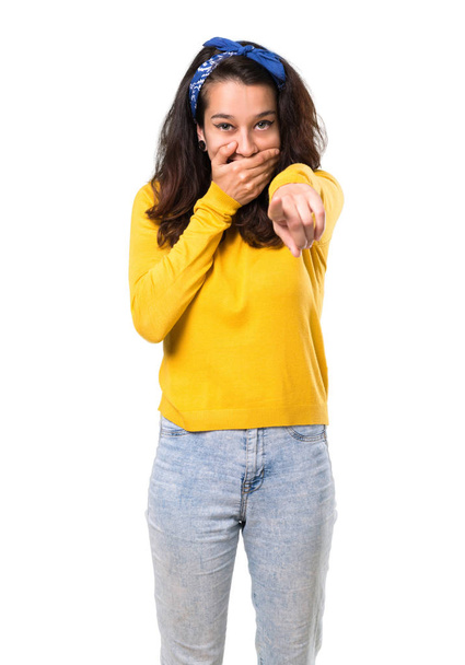 Young girl with yellow sweater and blue bandana on her head pointing with finger at someone and laughing a lot while covering mouth on isolated white background - Fotoğraf, Görsel