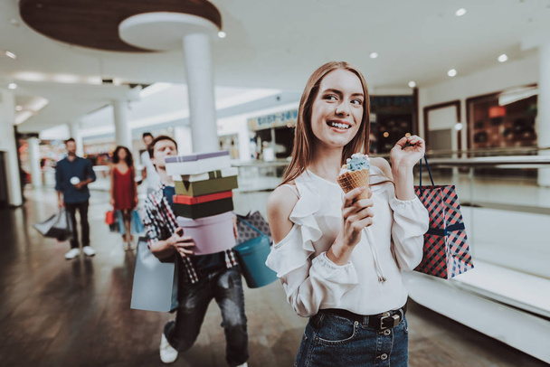 Carefree Woman with Ice Cream in Hand in Mall. Young Couple. Tired Man. Shopping Concept. Black Friday Concept. Happy Woman. Holding Packages. Pack from Mall in Hands. Ice Cream in Hand. - Фото, изображение