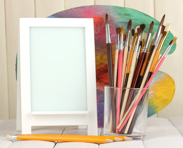 Photo frame as easel with artist's tools on wooden background - 写真・画像