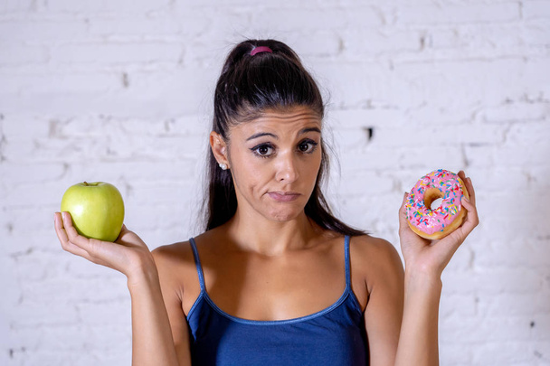 Beautiful young woman tempted having to make choice between apple and doughnut in healthy unhealthy food, detox eating, calories and diet concept. - Fotó, kép