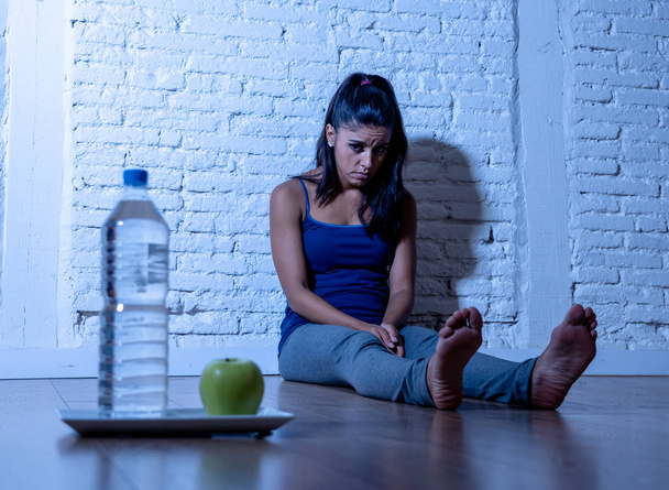 Desperate dieting young woman sitting on the ground looking sad at one apple fruit and water as symbol of crazy diet willpower and nutrition disorder. - Zdjęcie, obraz
