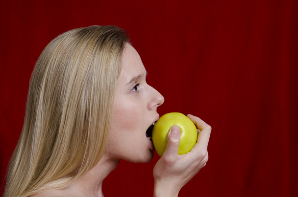 young blonde biting on apple on red background - Foto, Imagen