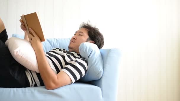 Ασιατική άνθρωπο διαβάζοντας το βιβλίο ξαπλωμένη στον καναπέ - Πλάνα, βίντεο