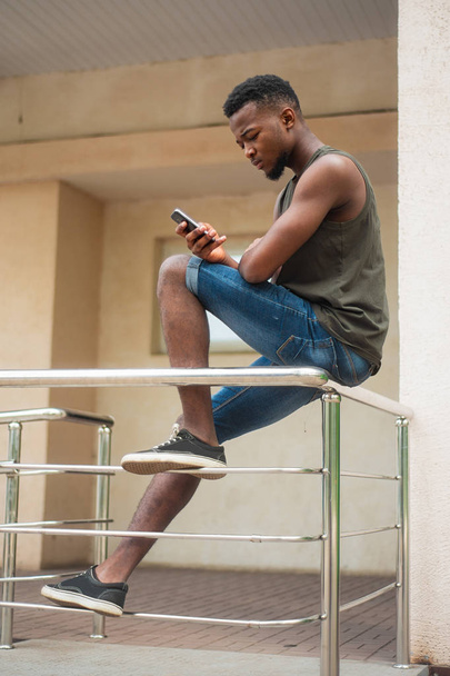 Concentrated urban hipster, a young man using a smartphone. African American teenager holding a mobile smartphone sitting on the railing - Valokuva, kuva