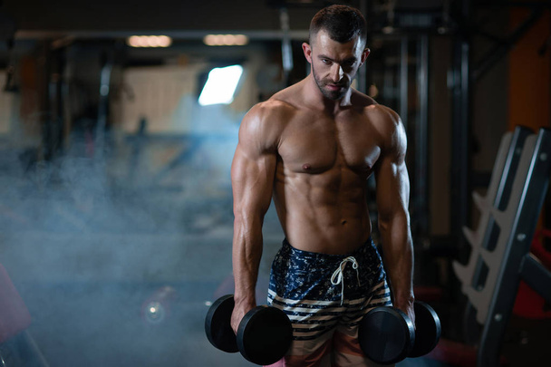 Beautiful young muscular man posing with dumbbells in hand at the gym - Photo, image