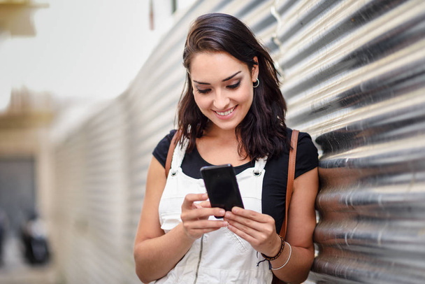 Chica sonriente usando con su teléfono inteligente al aire libre. Mujer joven feliz en el fondo urbano. Concepto tecnológico
. - Foto, imagen