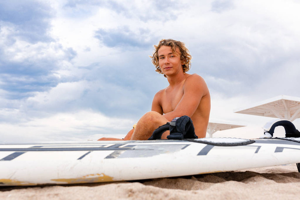 Komea mies istuu rannalla valkoinen tyhjä surffilauta odottaa aalto surffata paikalla meren rannalla. Käsite urheilu, kunto, vapaus, onnellisuus, uusi moderni elämä, hipster
. - Valokuva, kuva