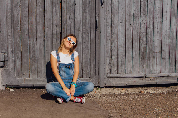 Portrait of a smiling woman dressed in overalls and sunglasses sitting on her skateboard next to the old wooden wall. - 写真・画像