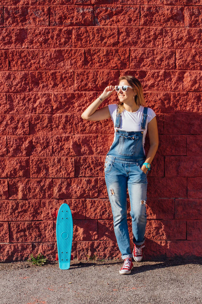 Portrait of a smiling woman dressed in overalls and sunglasses standing with her skateboard next to the red wall. - Fotoğraf, Görsel