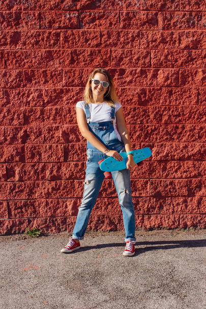 Portrait of a smiling woman dressed in overalls and sunglasses standing with her skateboard next to the red wall. - Zdjęcie, obraz