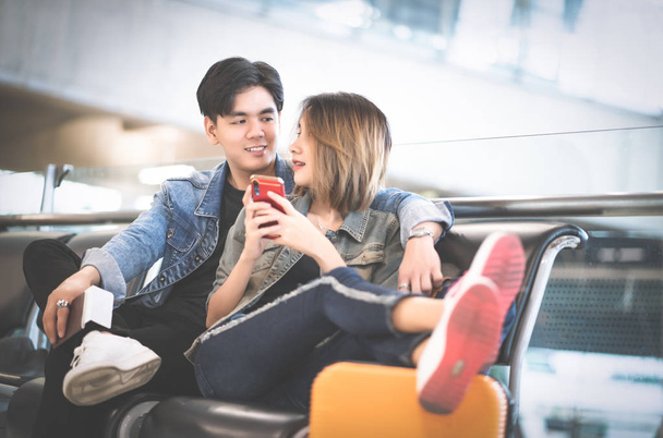 Young Asain loving couple in casual wear using smartphone while sitting in the airport terminal waiting for boarding. Teenager are traveling concept. - Foto, imagen