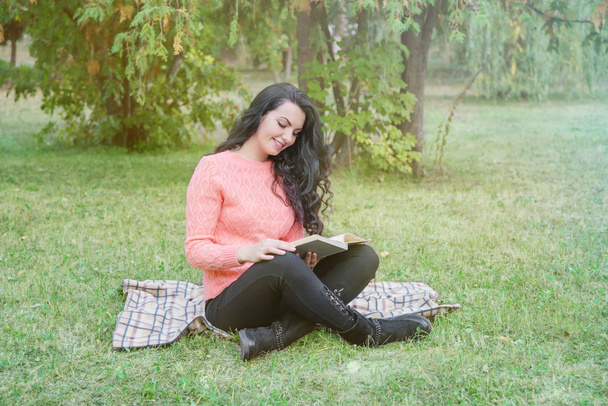 Beautiful long-haired brunette girl in a warm pink sweater sits in the park and reads a book - Photo, Image